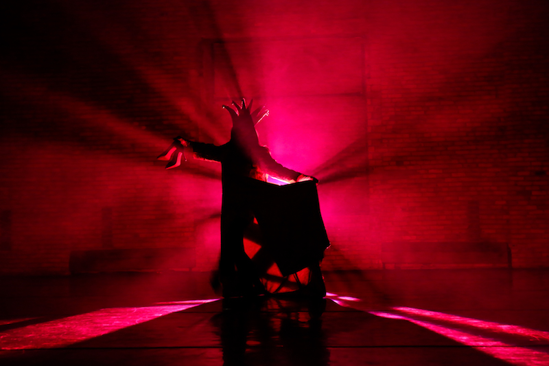 Stephen Petronio basked in redish, pink light. He wears a headpiece that obscures his face and vaguely looks like a crown. In one hand he holds out high heeled shoes. 